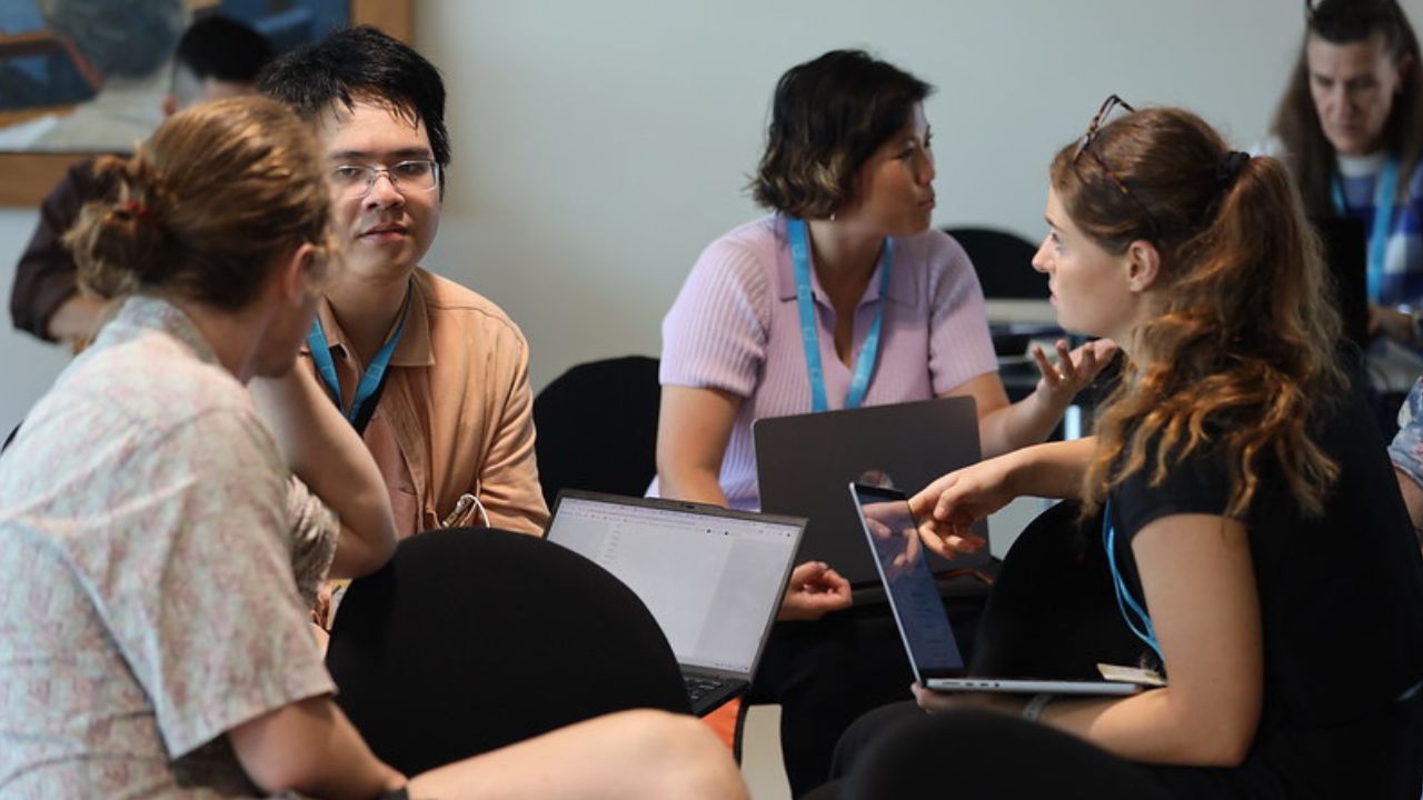 Group of 3 ADM+S researchers sitting at table with laptops with other members in the background