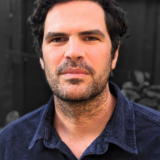 Photo_Mark_Doman.width-552 Photograph of Mark, a man with slightly wave, dark hair, brown eyes and stubble. Mark wears a dark blue, collared shirt.